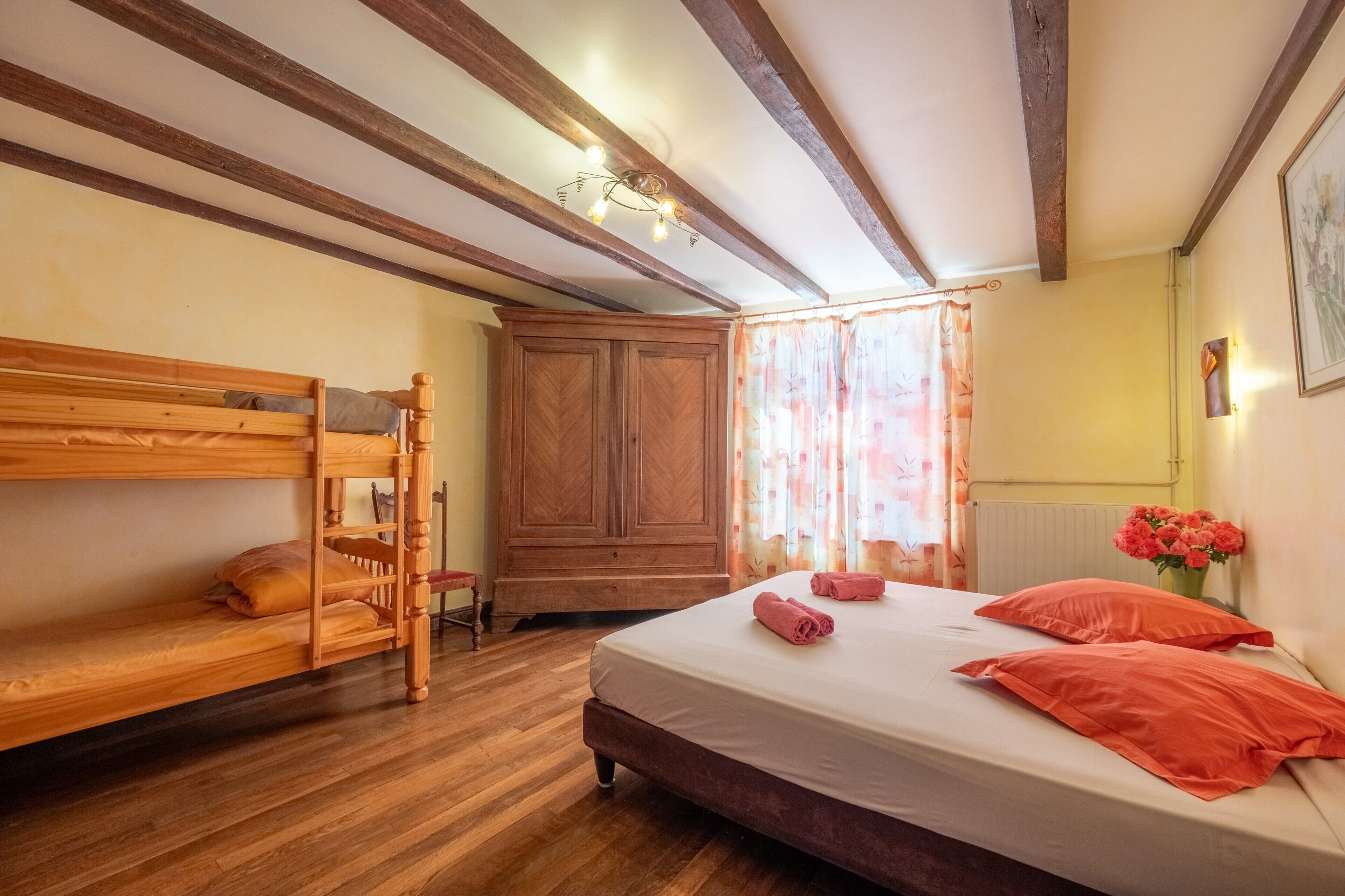 Une chambre avec un lit superposé, une grande armoire en bois, un lit avec des oreillers et une fenêtre avec des rideaux.