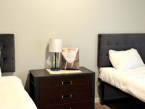 Une chambre d’hôtel avec une table de chevet en bois foncé, une lampe, un tableau encadré, un oreiller blanc et un lit avec une tête de lit grise.