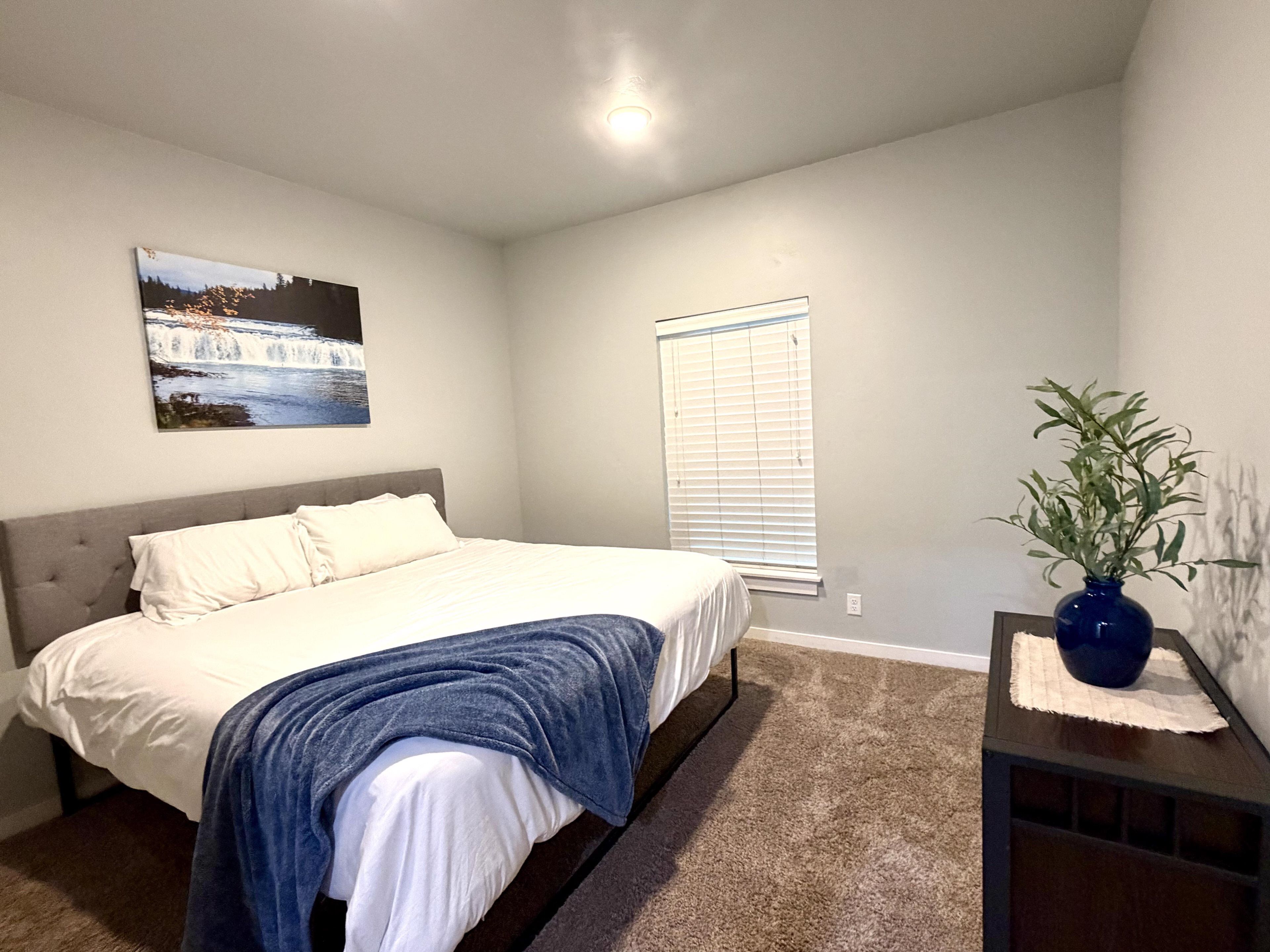 A bedroom with a bed, a window with blinds, a wall-mounted picture, and a small table with a vase.