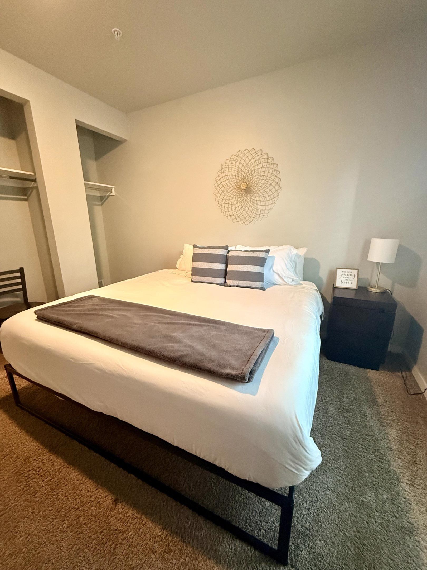 Una cama bien tendida con una manta marrón, una mesita de noche con una lámpara y una decoración en la pared.