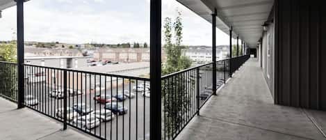 Un pasillo al aire libre cubierto, con suelo de hormigón, barandillas de metal y vistas a un aparcamiento y edificios al fondo.
