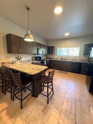 A kitchen with a granite countertop, dark cabinets, a central island with chairs, and a pendant light. - Brigham's Mill Retreat Between Grand Teton & Yellowstone National Parks (Rexburg)