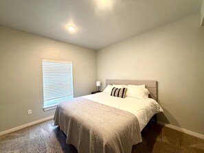 Un dormitorio con cama, mesita de noche, lámpara, ventana con persianas y luz de techo.