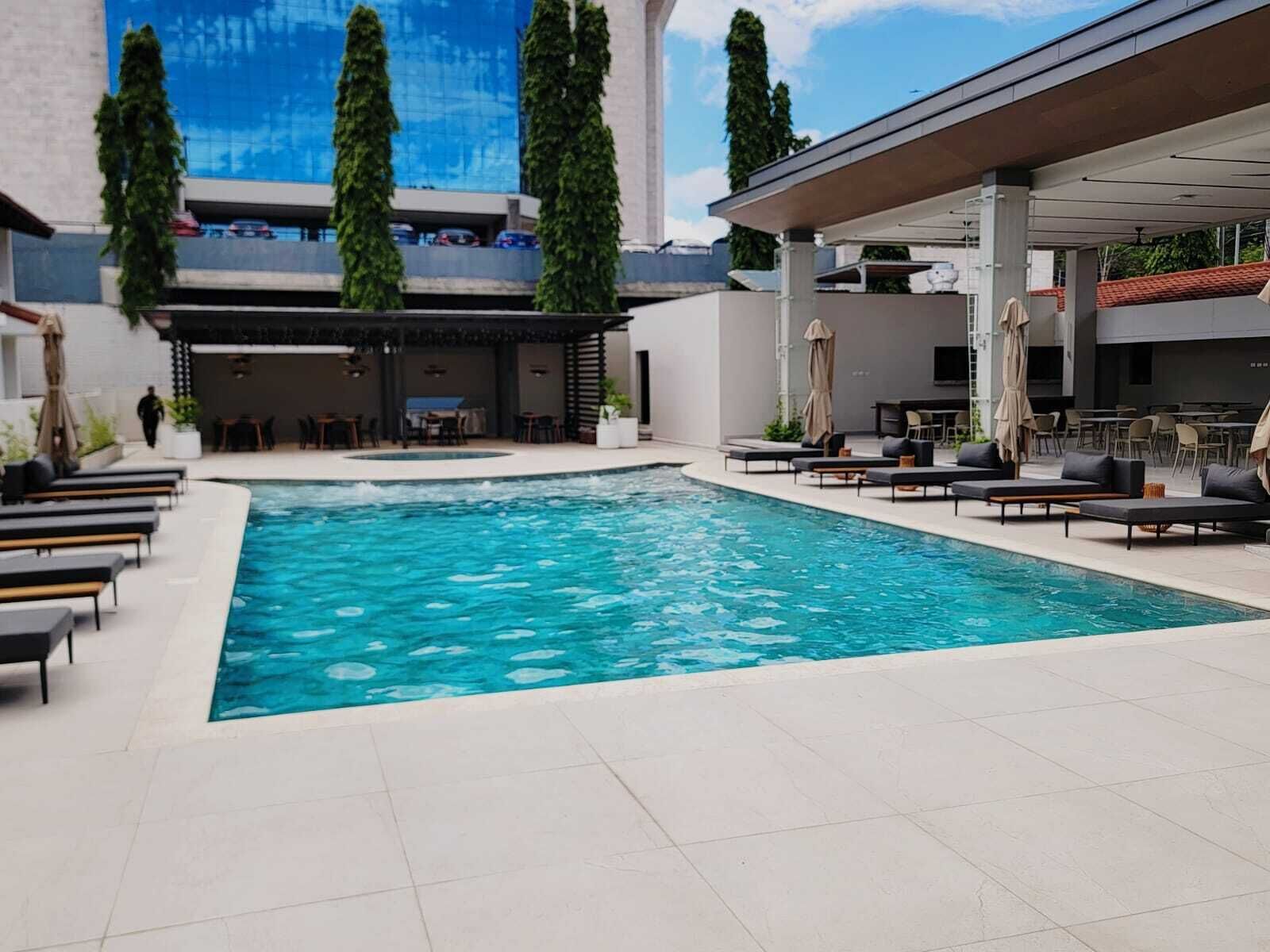 Une piscine entourée de chaises longues et de parasols, elle-même bordée d’un espace extérieur couvert comprenant des tables et des chaises.