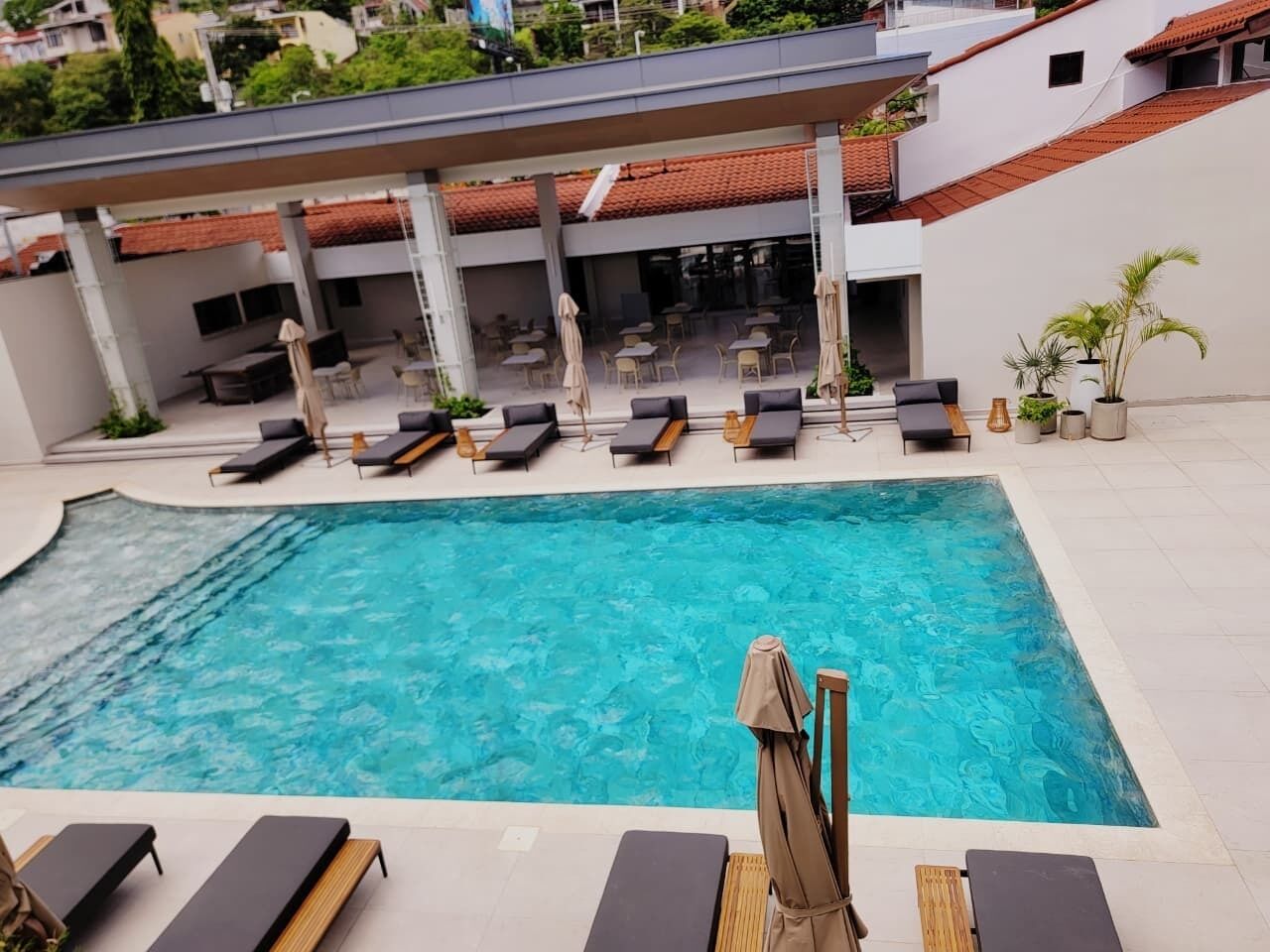 Una piscina con sillas de descanso y sombrillas, rodeada por un porche techado con mesas y sillas.