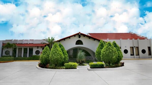 A building with a red-tiled roof, a central archway, and a paved courtyard with trimmed bushes. - Hotel Alameda (Tegucigalpa)