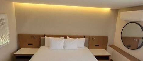 A hotel room with a large bed, bedside tables, a round mirror, and an air conditioning unit.