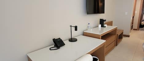Habitación de hotel con un escritorio blanco, una silla, una mesita, un televisor montado y un cesto de basura.