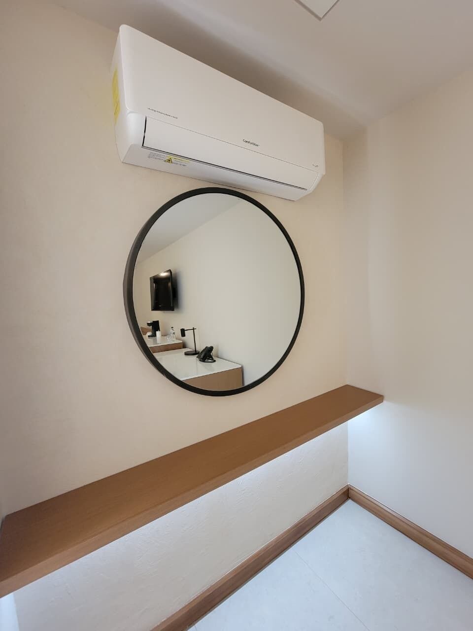 Un baño con un espejo redondo, un equipo de aire acondicionado blanco, un estante de madera y suelo de baldosas.