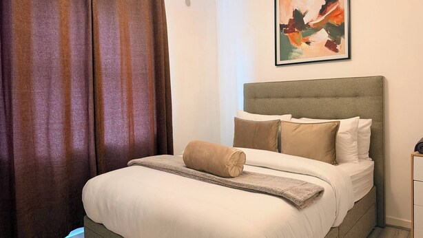 Un letto rifatto con cuscini e coperta, una testiera, un quadro appeso al muro e una tenda.