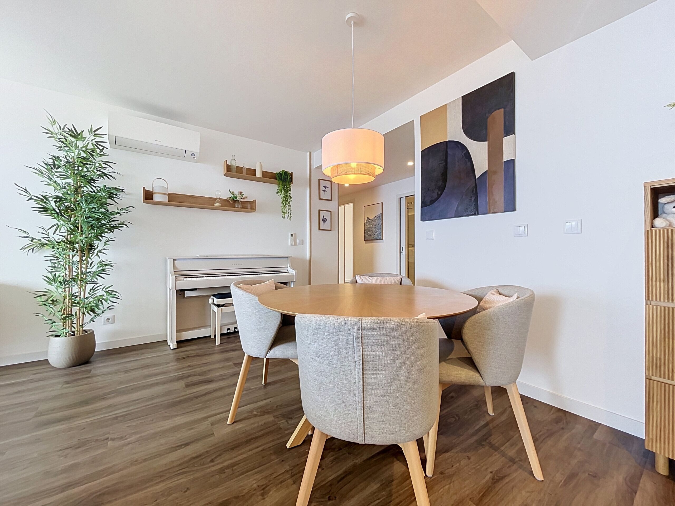 Ett modernt matområde med ett runt träbord, grå klädda stolar, en hängande lampa, ett piano och en stor, abstrakt målning på väggen.