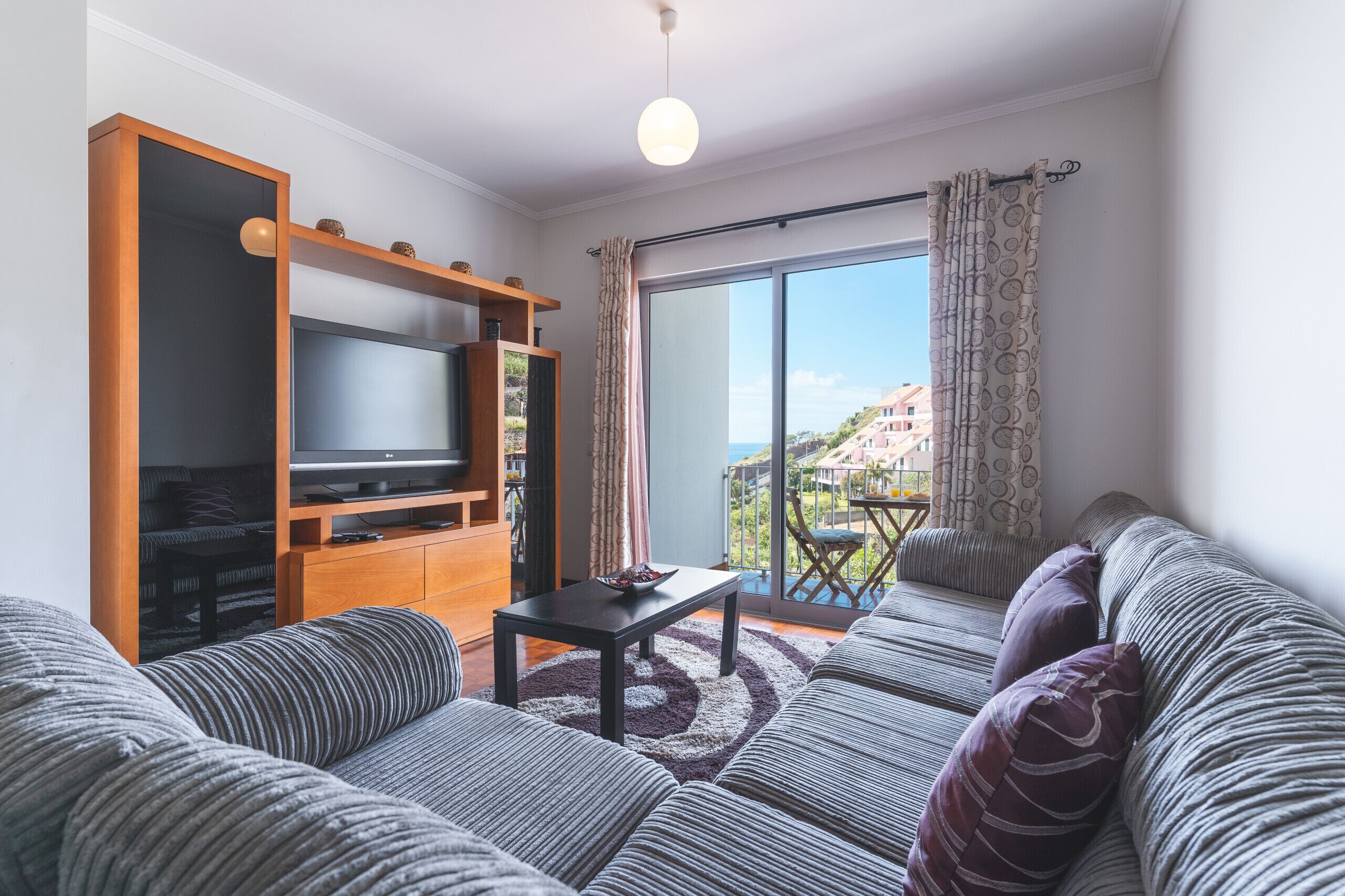 A living room with a sofa, a TV on a wooden stand, a balcony with a table, and a view of buildings.