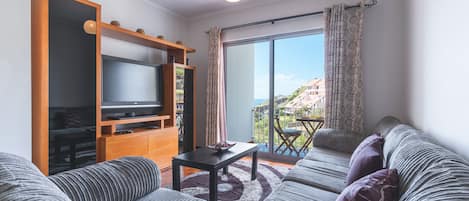 A living room with a sofa, a TV on a wooden stand, a balcony with a table, and a view of buildings.