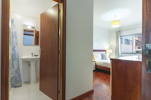 A hotel room with a wooden floor, a bed with a nightstand, a window with a view, and a bathroom with a sink and shower curtain. - Bella s Place Apartment by Atlantic Holiday (Santa Cruz)