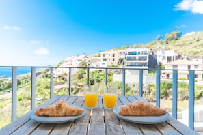 A balcony with two croissants and two glasses of orange juice on a wooden table, overlooking a coastal town. - Bella s Place Apartment by Atlantic Holiday (Santa Cruz)