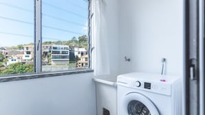 A compact laundry room with a washing machine, a small sink, and a window with a curtain. - Bella s Place Apartment by Atlantic Holiday (Santa Cruz)
