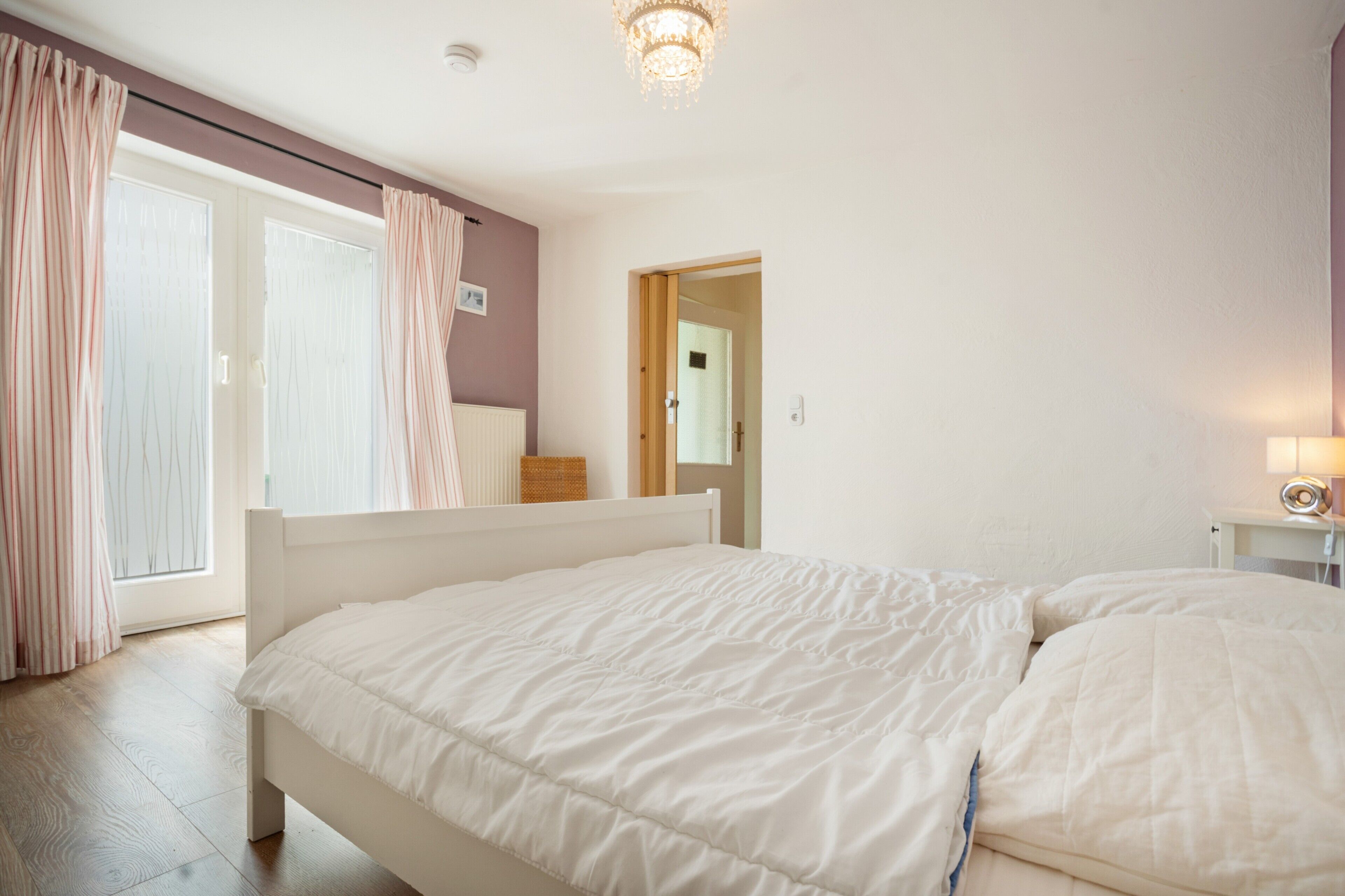 Ein Schlafzimmer mit einem Bett, einem Nachttisch, einer Lampe und Blick ins Freie durch ein Fenster.