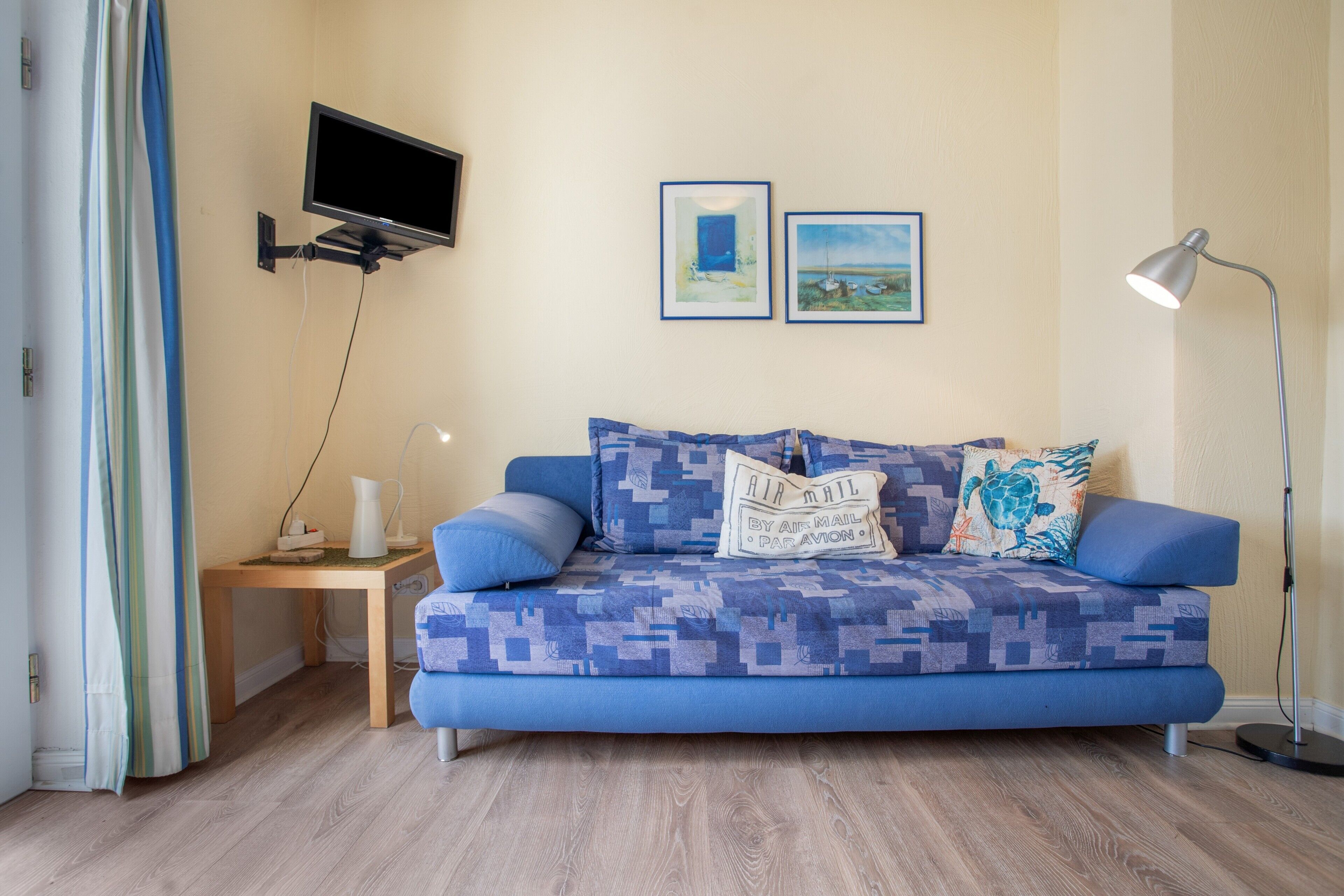 Ein Zimmer mit einem blauen Sofa, einem kleinen Beistelltisch, einer Stehlampe und zwei gerahmten Bildern an der Wand.