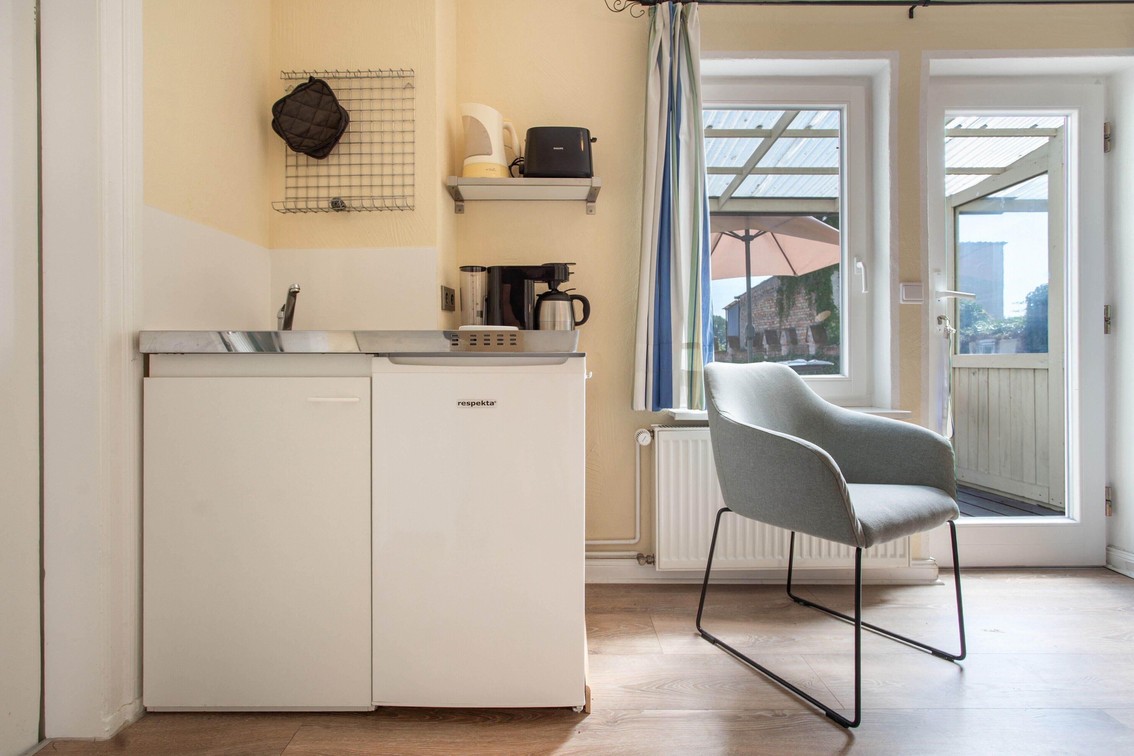 A compact kitchen with a dishwasher, a coffee maker, and a kettle on a shelf. There is a chair in front of a window with a view of a building outside.