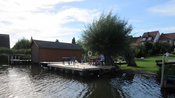 A dock with a table and chairs, a lifebuoy, and a building in the background. - Apartment 'Fewo Schwan' with Shared Terrace, Shared Garden and Wi-Fi (Röbel)