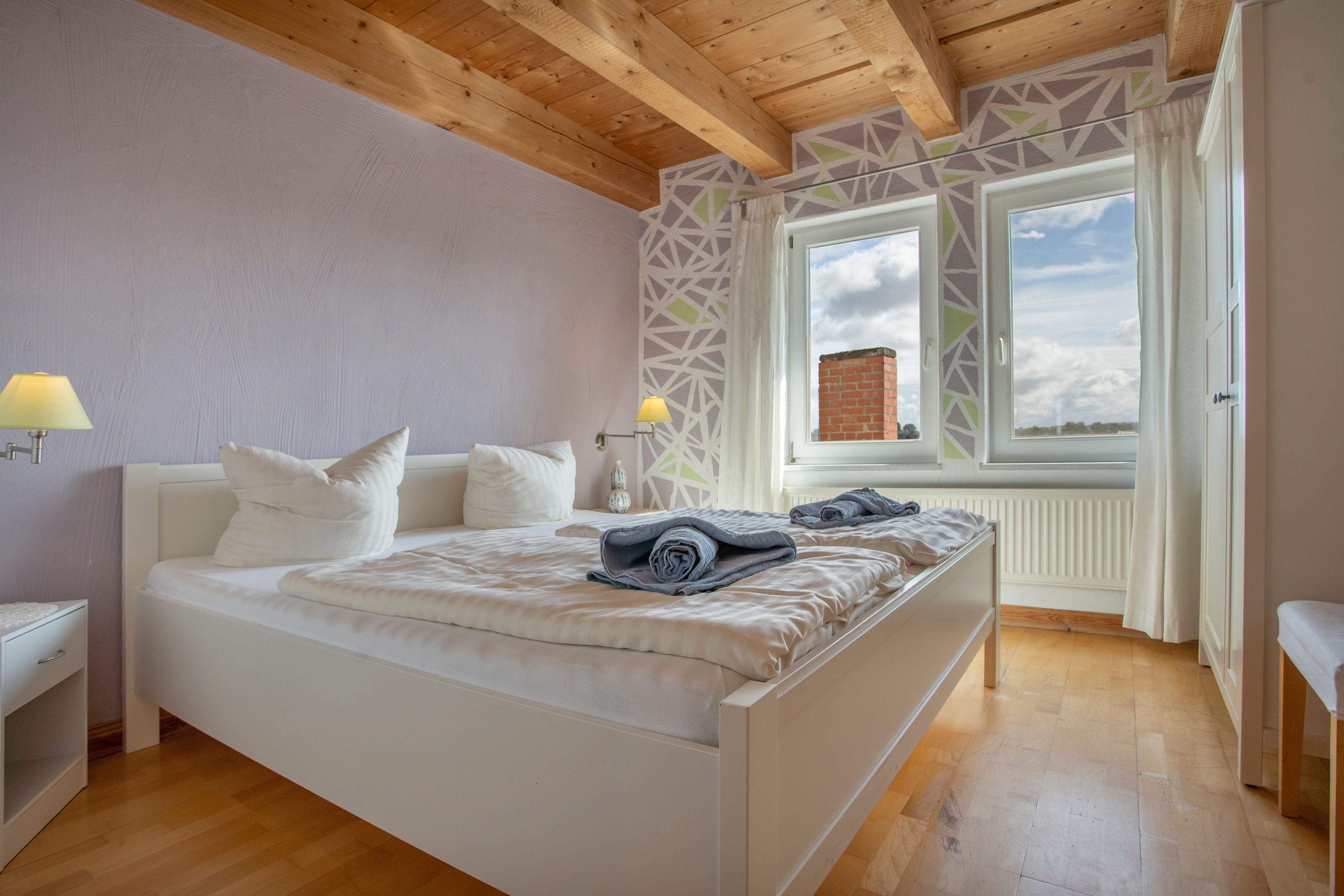 Ett sovrum med ett träpanelat tak, en säng med vita sängkläder, två sänglampor och ett fönster med utsikt över en byggnad med tegelstensfasad.
