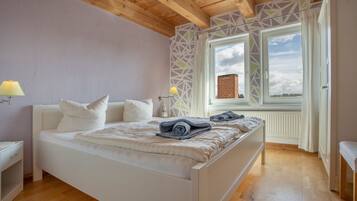 Ett sovrum med ett träpanelat tak, en säng med vita sängkläder, två sänglampor och ett fönster med utsikt över en byggnad med tegelstensfasad.