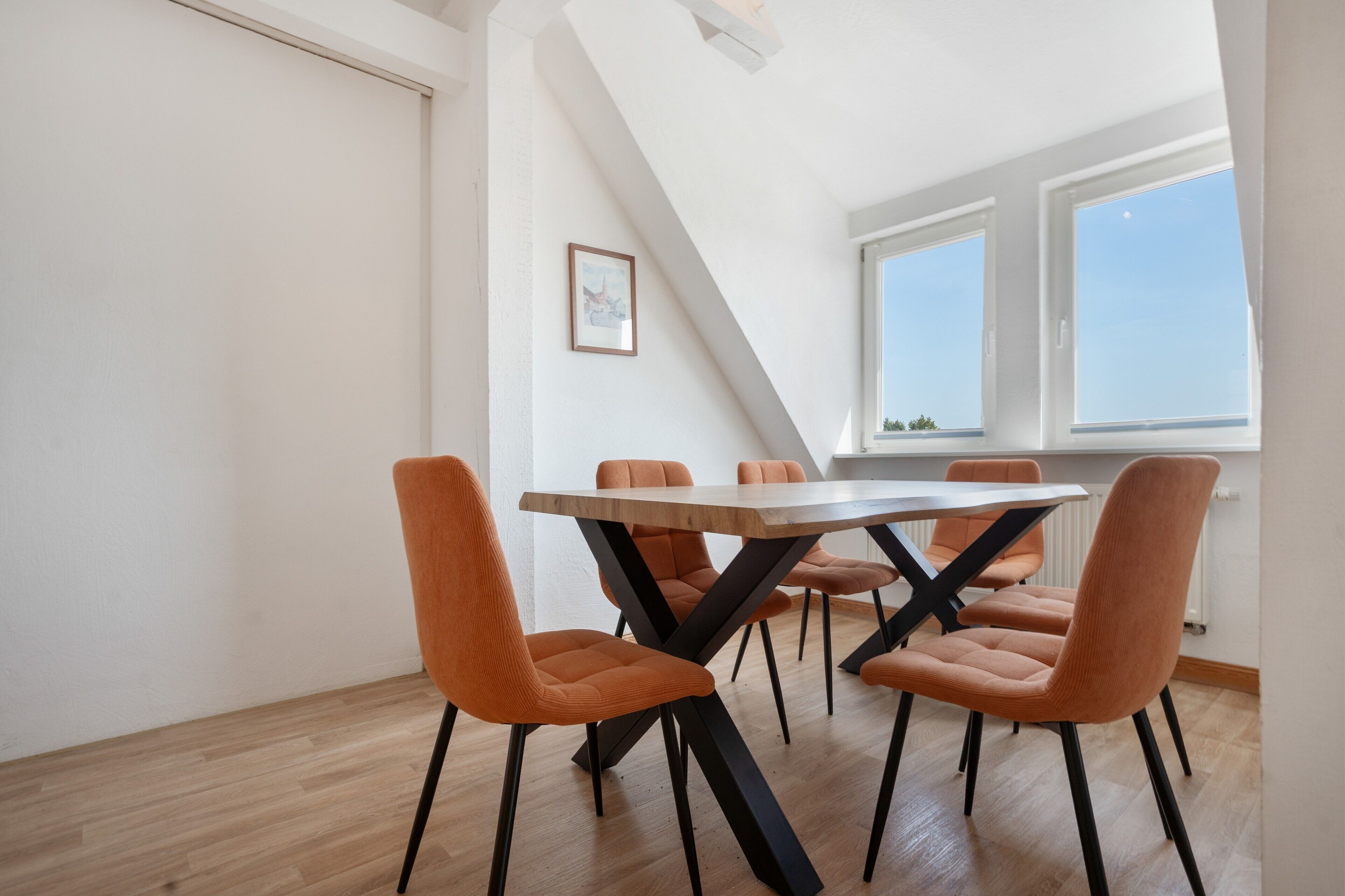 Ein Essbereich mit einem Holztisch und orangefarbenen Stühlen, einem gerahmten Bild an der Wand und Blick auf den Himmel durch ein Fenster.