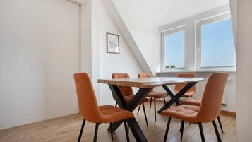Ein Essbereich mit einem Holztisch und orangefarbenen Stühlen, einem gerahmten Bild an der Wand und Blick auf den Himmel durch ein Fenster.