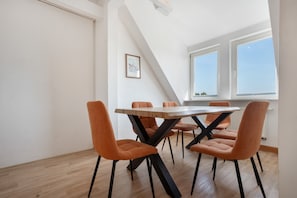Ein Essbereich mit einem Holztisch und orangefarbenen Stühlen, einem gerahmten Bild an der Wand und Blick auf den Himmel durch ein Fenster.