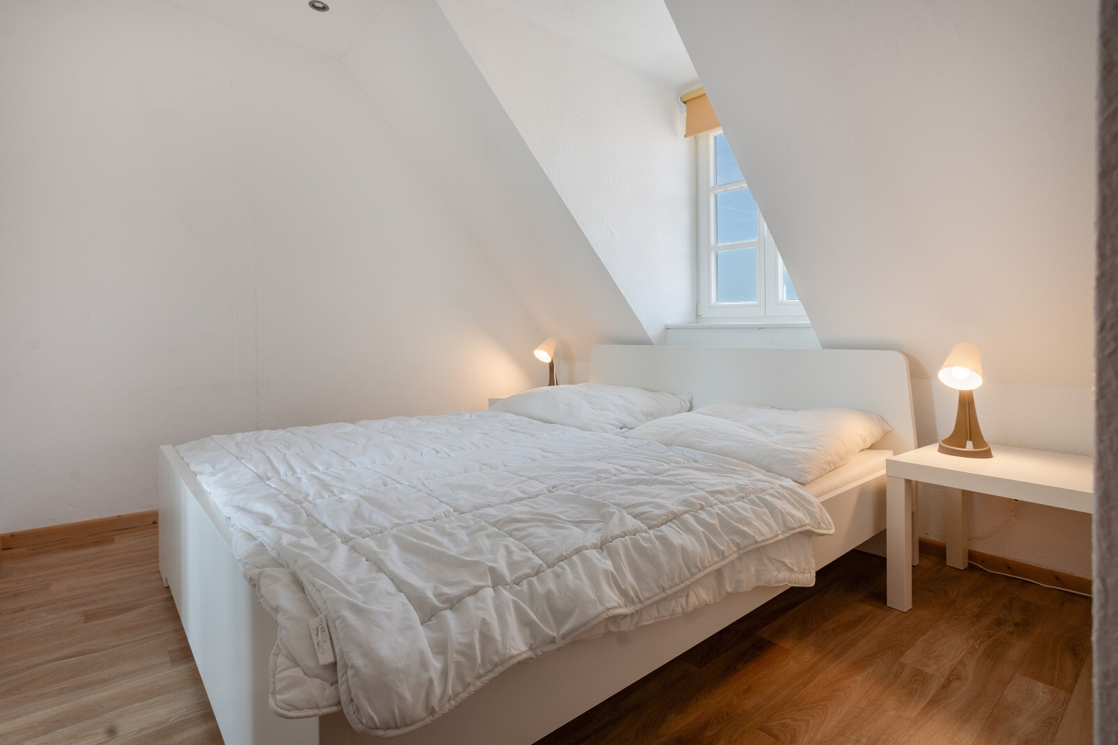 Une chambre avec un plafond en pente, un lit avec une literie blanche, deux lampes de chevet et une fenêtre donnant sur un beau paysage.