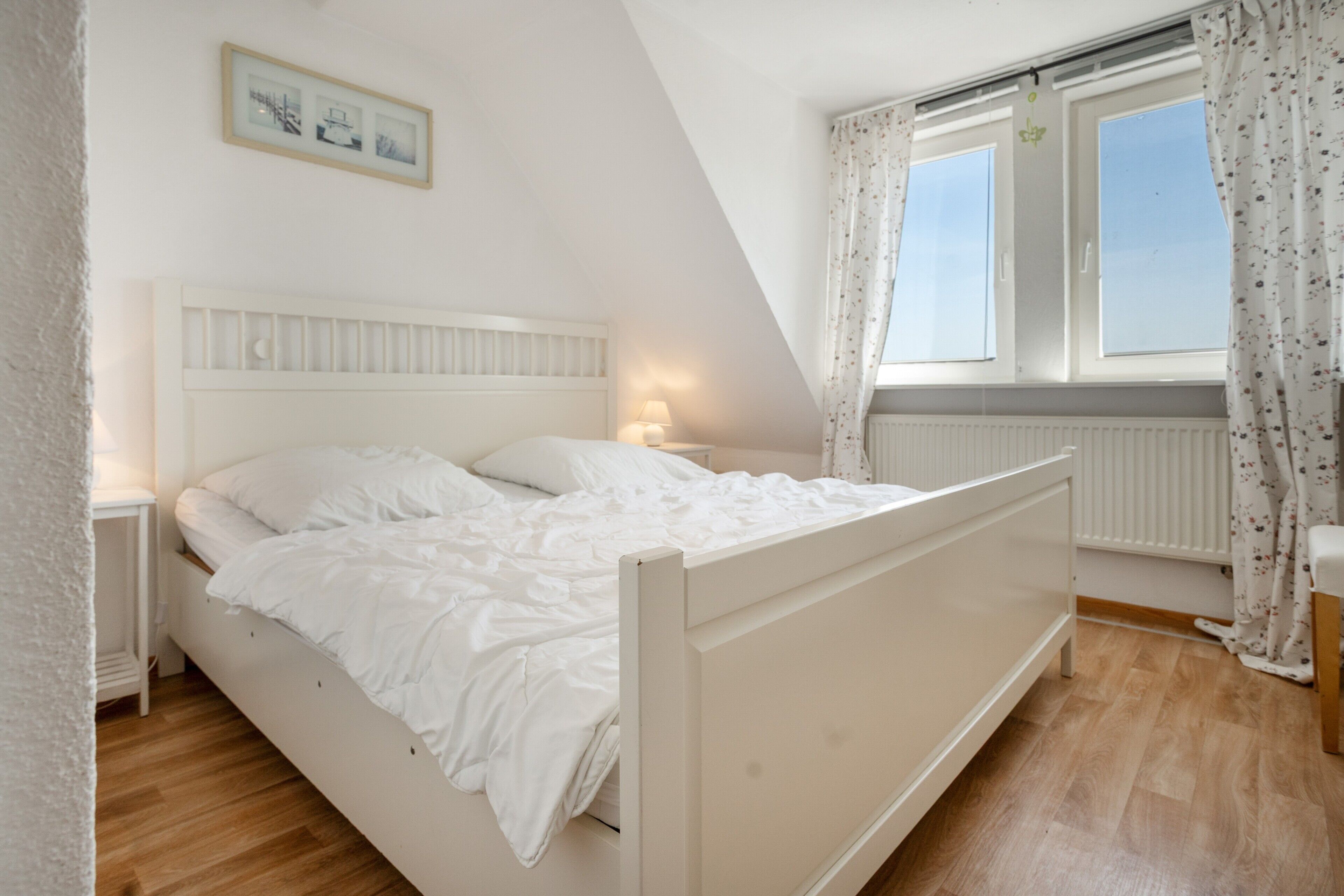 A bedroom with a single bed, a nightstand, a window with curtains, and a wall-mounted picture.