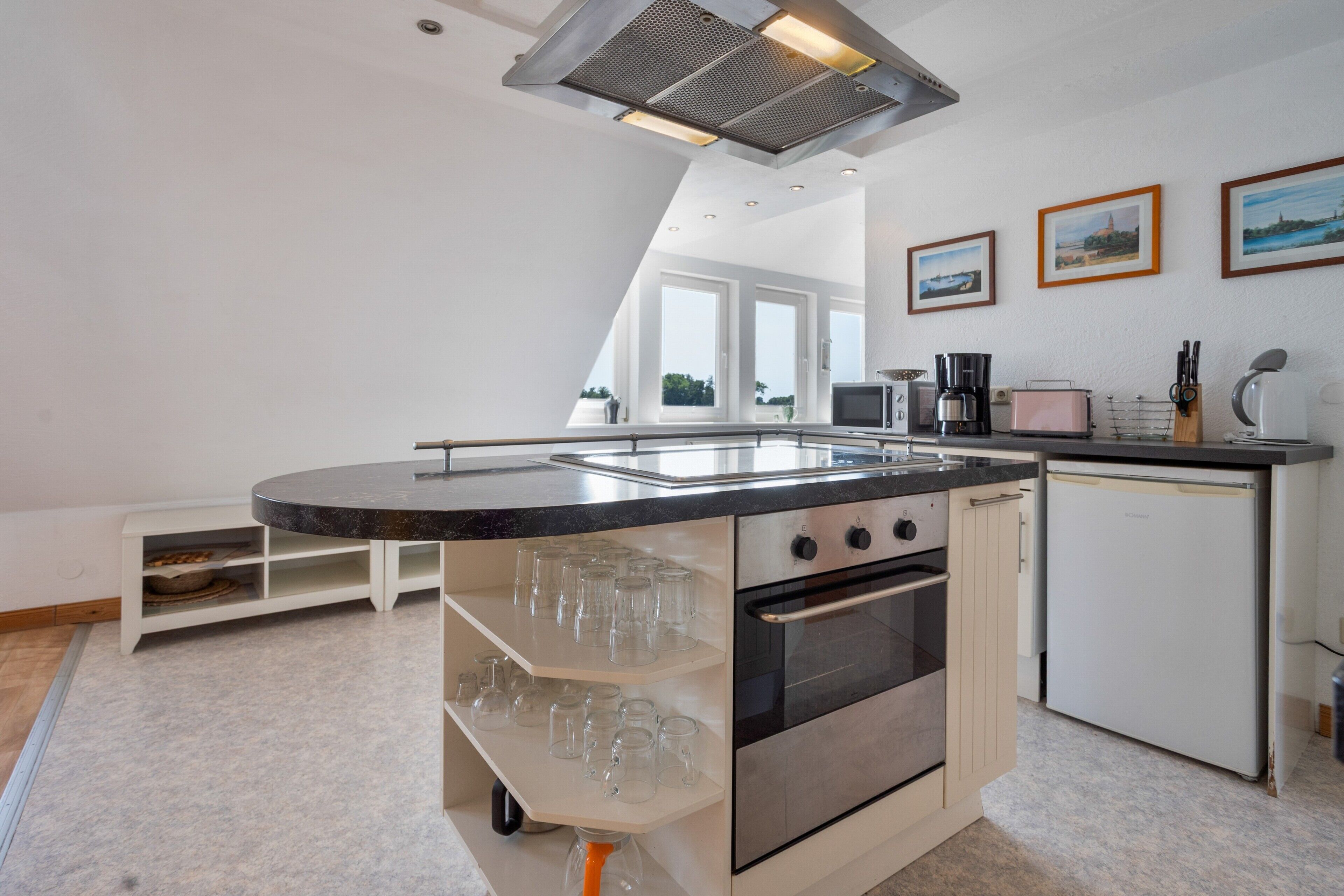 Una cocina moderna con una isla central, horno empotrado y microondas. Hay cuadros enmarcados en la pared y un ventanal que proporciona luz natural.