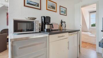 A compact kitchen with a microwave, coffee maker, and toaster. There are cabinets and a countertop. A bed is visible in the adjacent room.