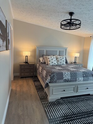 A bedroom with a bed, two bedside tables, a floor lamp, a ceiling fan, and a patterned rug.