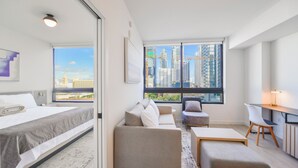 Una habitación de hotel moderna con cama, sofá, una mesita y un escritorio. Amplias ventanas ofrecen vistas a un paisaje urbano.