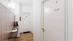 Un corridoio con pavimento in legno, una porta bianca con una stella e un testo, una lampada a parete e una piccola mensola con un bollitore e una bilancia.