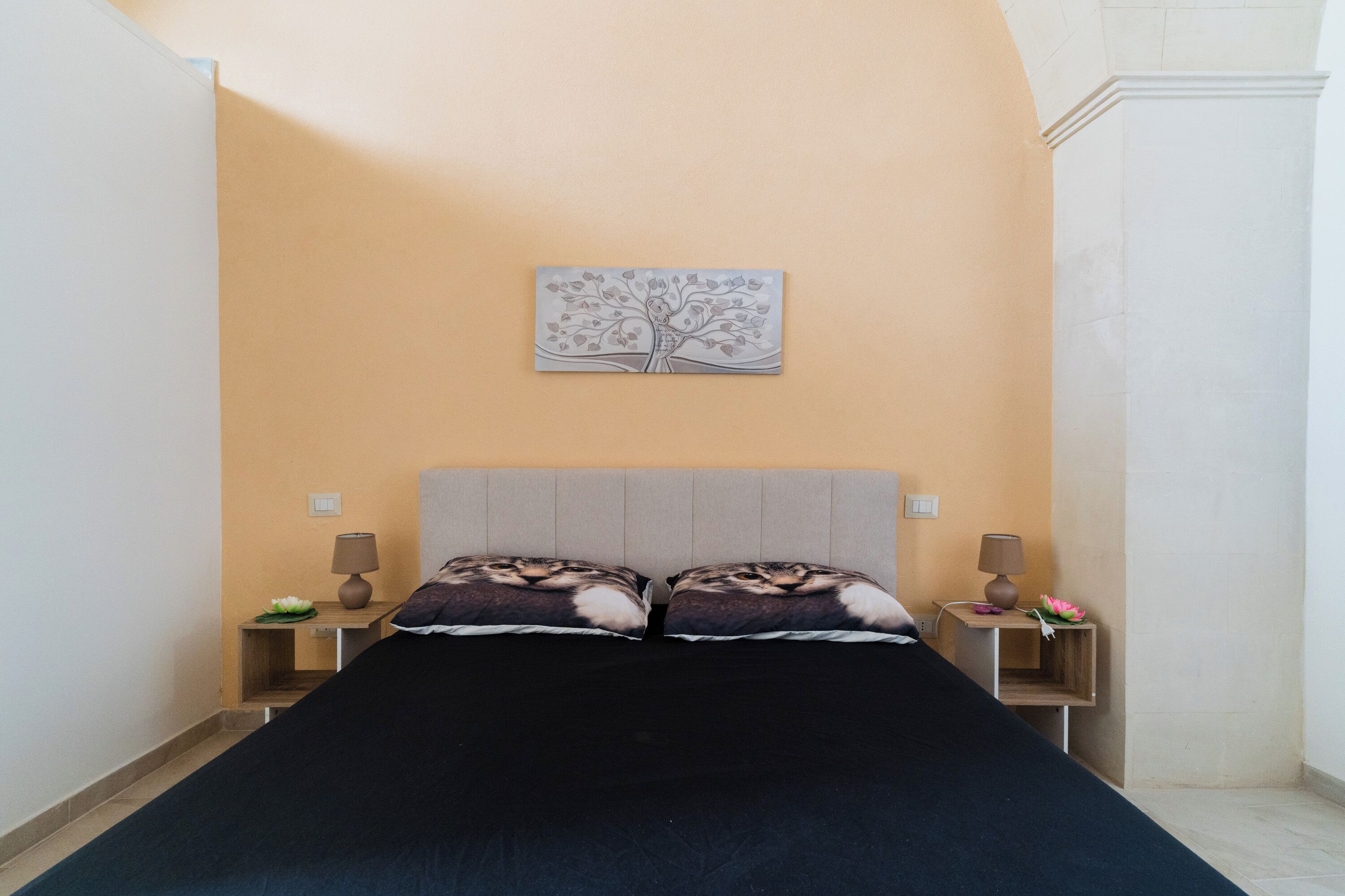 A bedroom with a bed, two nightstands, a lamp on each, a wall art piece, and a small table with a flower.
