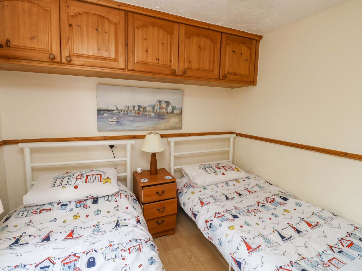 Une chambre avec deux lits simples, une commode en bois, une table de chevet, une lampe et un tableau représentant une ville côtière.