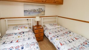 Un dormitorio con dos camas individuales, un aparador de madera, una mesita de noche, una lámpara y un cuadro de un pueblo costero.
