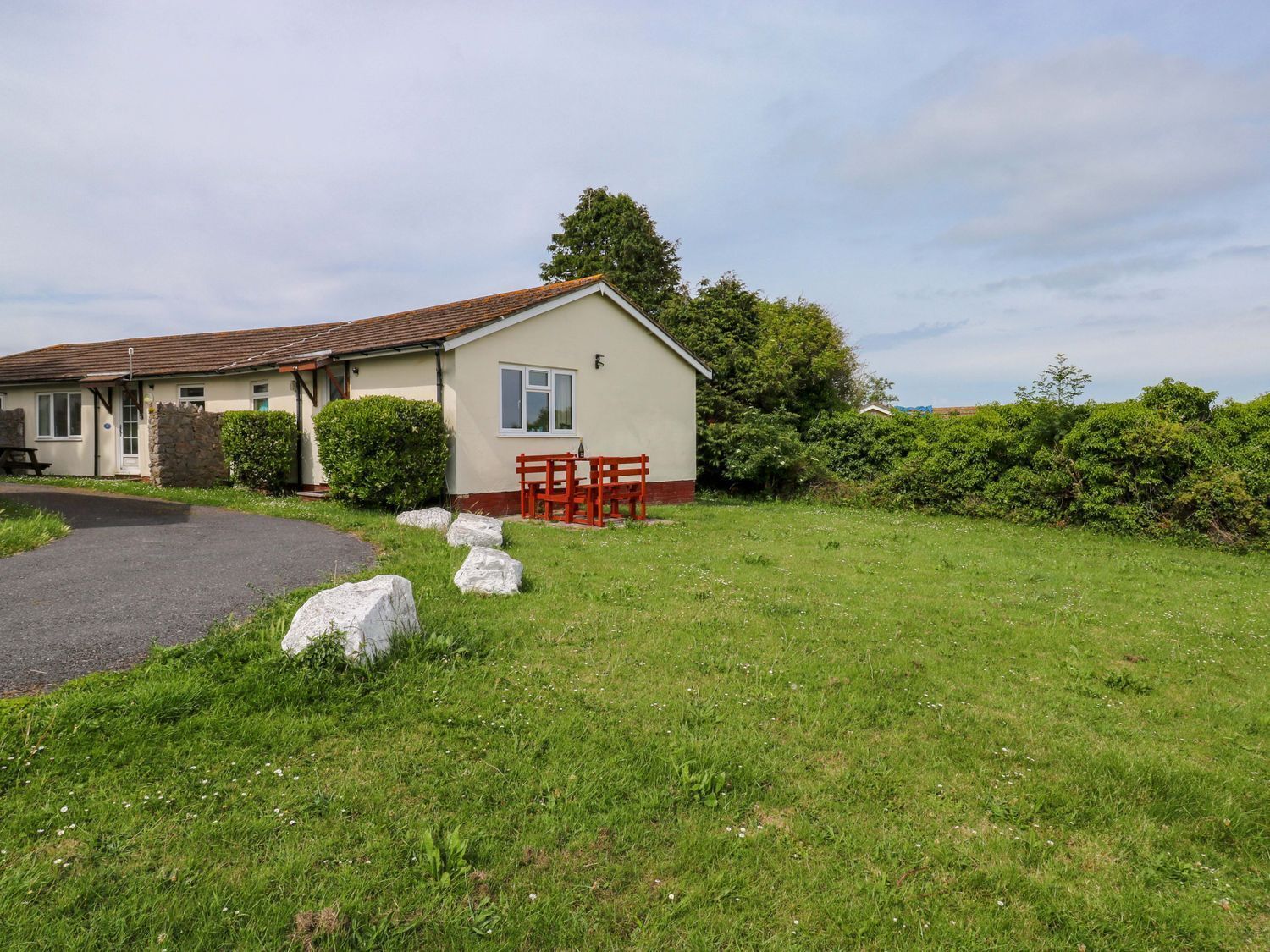 A single-story house with a paved driveway, a small patio with red furniture, and a well-maintained lawn.