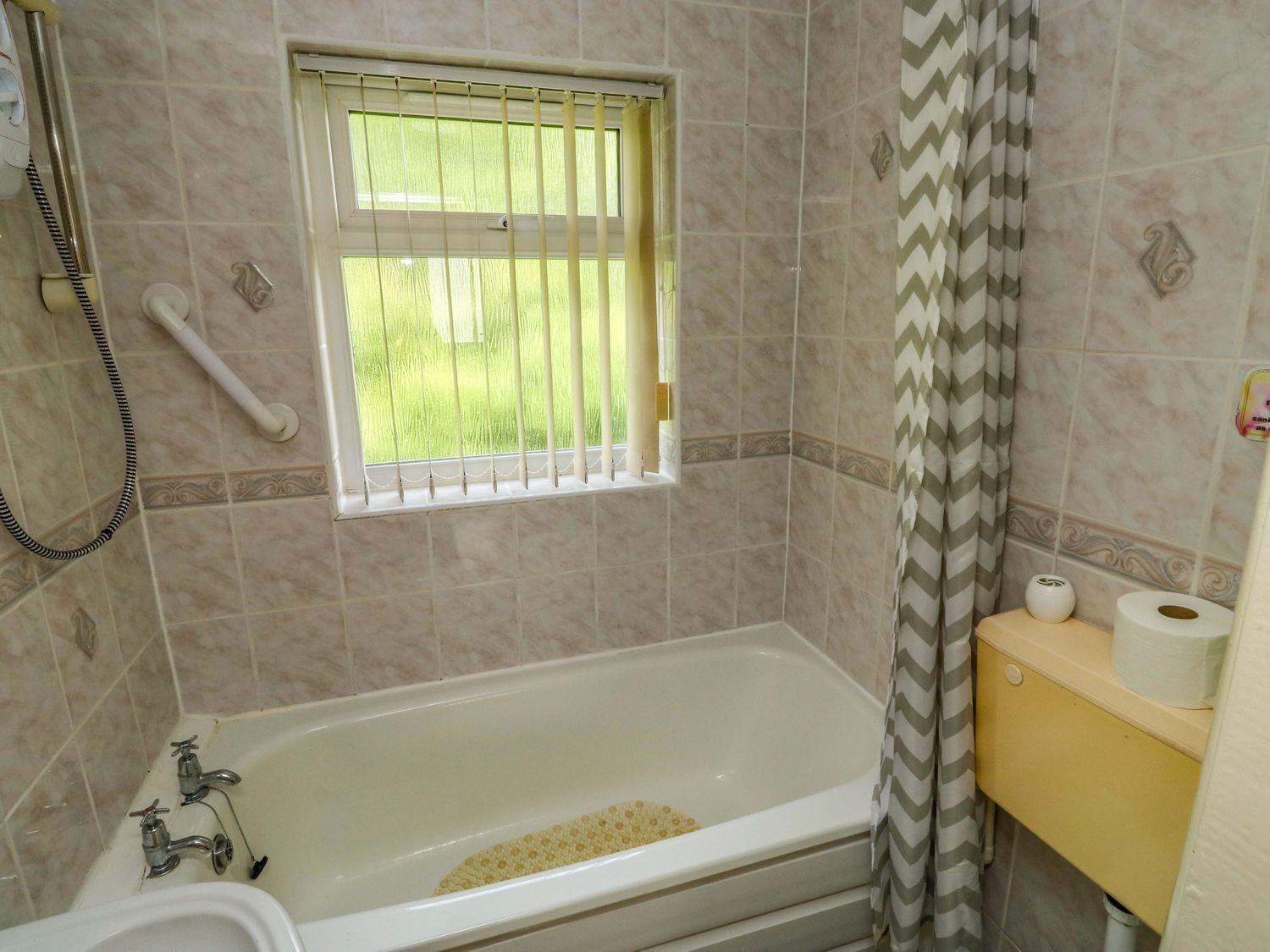 A bathroom with a bathtub, a window with bars, a shower hose, and a toilet with toilet paper.