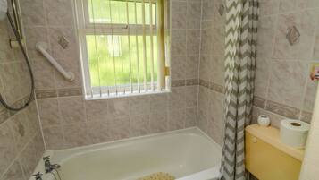 A bathroom with a bathtub, a window with bars, a shower hose, and a toilet with toilet paper.
