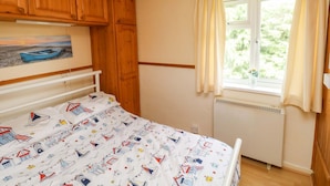 A bedroom with a bed, wooden cabinets, a painting, a window with curtains, and a radiator. - Seascape Retreat (Brixham)