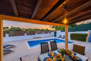 A poolside dining area with a view of a swimming pool, outdoor seating, and a covered patio.