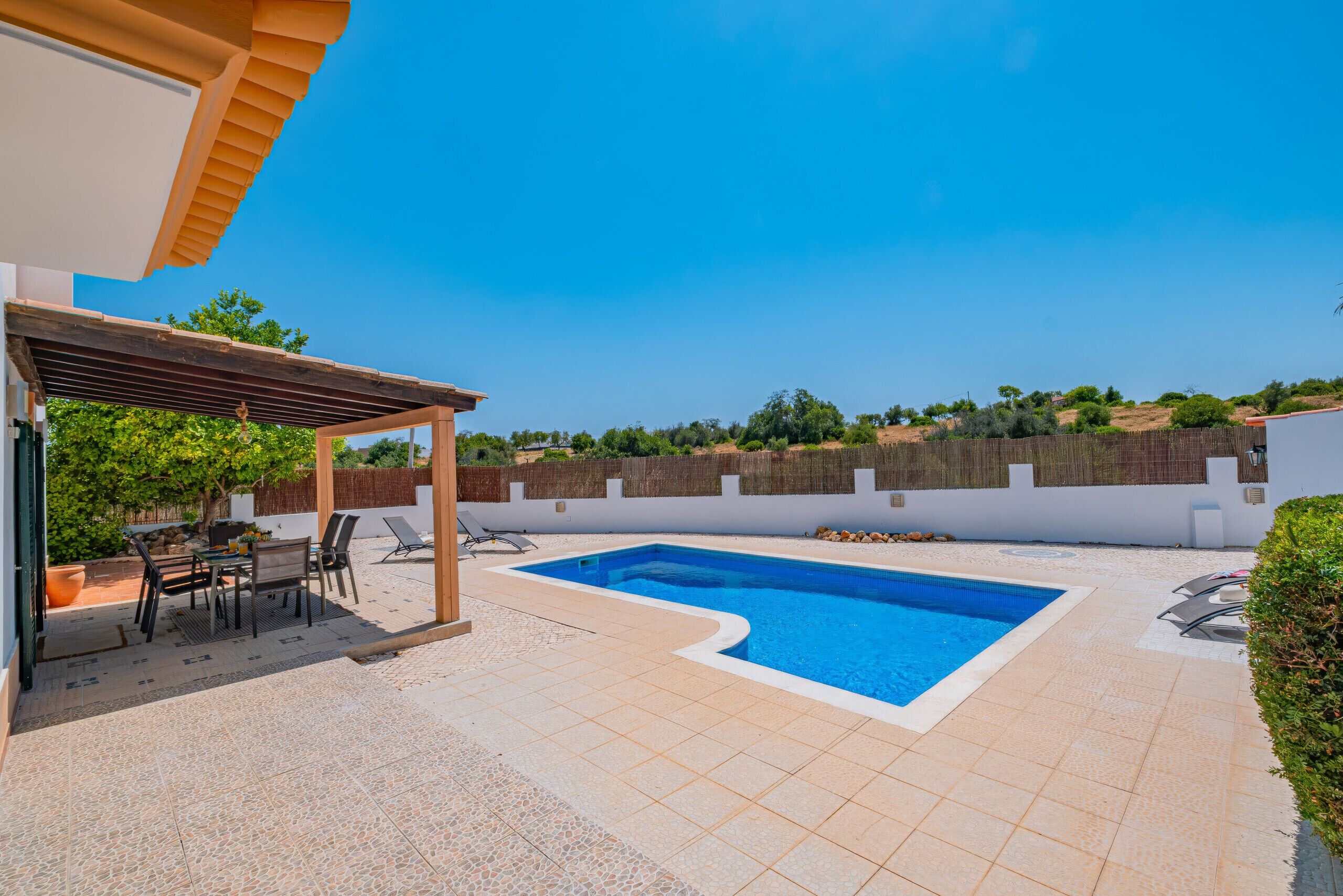 Une piscine agrémentée d’une pergola et d’un espace détente, entourée d’un mur blanc et bordée d’un paysage vallonné.