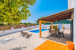A sunny outdoor patio with a swimming pool, seating area, and a pergola.