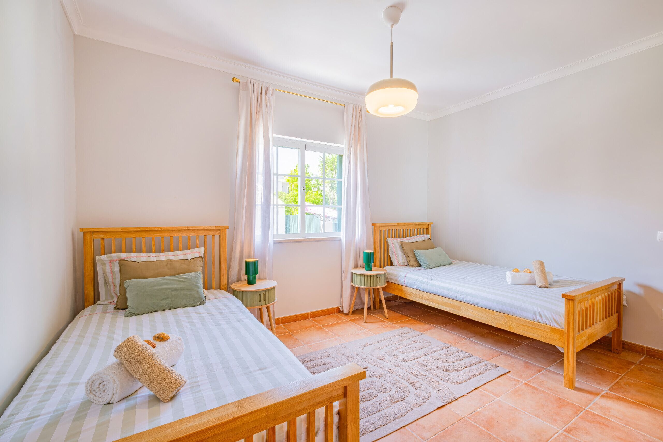 Un dormitorio con dos camas de madera, una ventana con cortinas, una mesita con lámpara y una lámpara colgante.