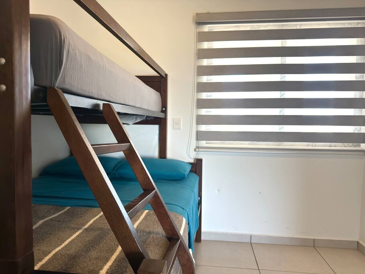 Un lit superposé avec une échelle, un lit simple avec un matelas bleu et une fenêtre avec des stores horizontaux.