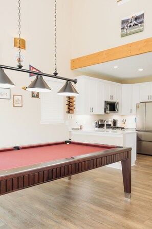 Une table de billard dans une cuisine moderne, avec des meubles blancs et un réfrigérateur en acier inoxydable.