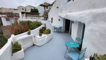 Un patio estrecho con paredes blancas, una pequeña mesa con sillas y plantas en macetas.